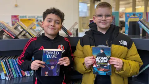Two boys - one with dark hair holding a purple book and another with light hair and glasses - he is holding a blue book that says Star Wars