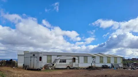 West Mercia Police A wide steel frame building with white and green panels. A caravan is sat in front. Blue skies and clouds can be seen in the background.