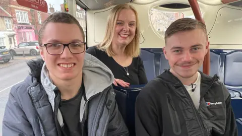 BBC/RICHARD EDWARDS Tyler and Alfie siting next to each other on a bus. Tyler is wearing glasses and a grey coat. Alfie is wearing a black hoodie and a white T-shirt. Behind them is Lauren Dent-McShane, who has blonde, shoulder length hair. She is wearing a black top and smiling at the camera.