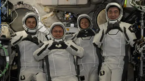 Four astronauts wearing white spacesuits and helmets pose for cameras smiling and making heart shapes with their hands and fingers. They are surrounded by wires and other equipment inside the International Space Station.