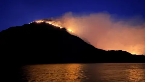 A fire runs down a hill, with smoke billowing to the right hand side. The orange of the flames and smoke is reflected in a lake at the bottom of the hill. 