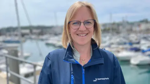 Jenny Giles standing in front of Marina Qeii. She wears a Blue Guernsey Ports jacket, has blond hair of neck length and wears glasses.