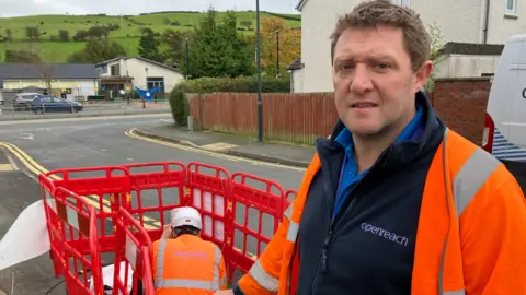 Tegryd Rees está vestindo uniforme Openreach e colete de alta visibilidade, com um colega trabalhando ao fundo