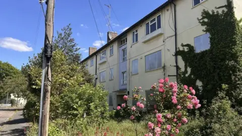 A three-storey block of flats, with boarded up windows on the first two floors. It has trees and shrubbery out in front of the building.