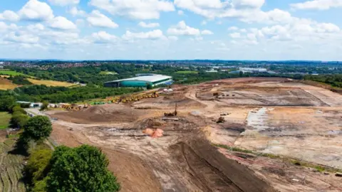 Image shows a large swathe of land that has been taken back to soil level which is surrounded by trees and fields. There are tyre and caterpillar tracks crisscrossing the land, which looks like the beginning of a building site.