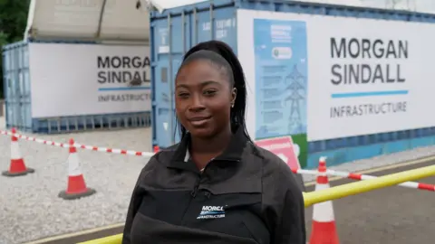 Hannah Karpel/BBC A young woman  in a black Morgan Sindall Infrastructure zip-up jacket standing in the foreground at a construction or infrastructure site. Behind the person are two blue site cabins or container units displaying large Morgan Sindall Infrastructure signs. Red-and-white traffic cones and barrier tape mark off a gravel area, with a yellow safety barrier running across the lower right of the image. Additional site signage is attached to one container, and electricity pylons are visible in the background.