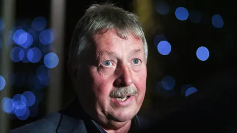 Reuters Sammy Wilson with spotlights successful  the background. He has abbreviated  grey-ish hair, bluish  eyes and a moustache and wearing a acheronian  grey blazer. 