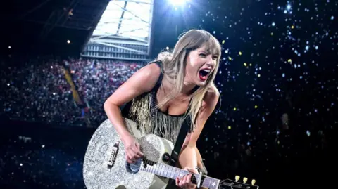 Getty Images Singer Taylor Swift performs with a sparking guitar in front of a huge crowd. She is smiling and wearing red lipstick