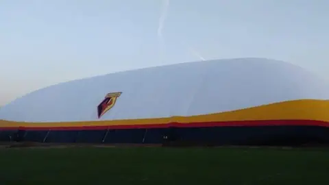 CovAir/Savills/Hertsmere Borough Council A large inflatable dome structure in Watford football club colours—white with a yellow and red band—stands on a grassy field. The Watford crest is visible on the side, and a clear sky stretches above the dome.