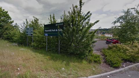 Signs for Thornhill Park and Ride from London Road and a red motorcycle on the right of the picture that can be seen in a gap in a hedge 