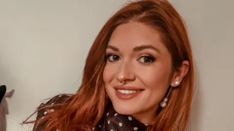 A selfie of a young woman with long red hair. She is wearing a brown silk dress with white polka dots and has tattoos on both arms. 