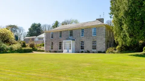 The picture shows a large Georgian country house built from pale stone, set back behind a wide, neatly cut green lawn. The house is symmetrical and two storeys high, with a central white front door and evenly spaced sash windows on either side and above. The roof is shallow and slate‑coloured, with chimneys visible at either end. In the foreground, the lawn stretches right up to the front of the building