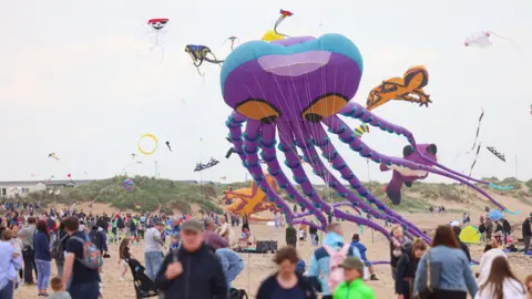 A large kite in the shape of an octopus being flown on Redcar beach. It is purple with large orange eyes. A number of smaller kites are also being flown in the area with dozens of people looking on.