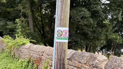 A poster reading SUPPORT OUR OBJECTON OFFERTON LANE DOES NOT WANT PADEL is attached to wooden lamp post near a stone fence in Offerton.
