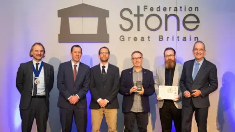 Salisbury Cathedral Six middle-aged men dressed smartly in suits, one holding an engraved award and another, a certificate, in front of a wall with 'Federation Stone, Great Britain' written on it. 