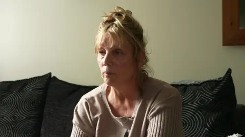 BBC A woman sat on her sofa looking away from the camera rather upset. She has blonde hair and a beige sweater on. Her sofa has a few black cushions. It's pushed up against a clear white wall with a mirror to the right hand side.