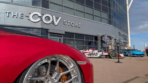 Super cars outside a grey building. Nearest to camera is a bright red car with a shiny wheel while a while HAAS car with pink livery can be seen in background. The building has The Cov Store written on it