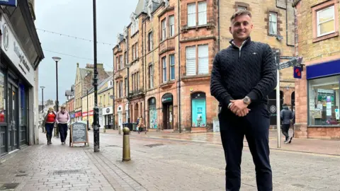 BBC A man in a dark top and dark trousers stands across the street from a large sandstone building