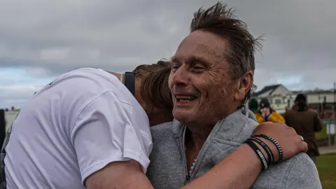 The Hull Boy Two men embrace. One has shoulder-length hair and is wearing a white T-shirt and three black wristbands, and the other has shorter hair and is wearing a grey top.