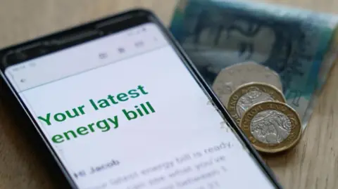 A phone with an email reading "your latest energy bill" on the screen, sat on a wooden table next to a greeny-blue five pound note, two pound coins and a 50p coin.