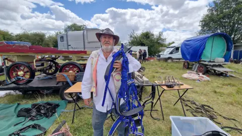 Man in a hat, waistcoat, shirt and jeans holding a blue harness. There are harnesses, carts, vehicles and people behind him on grass.