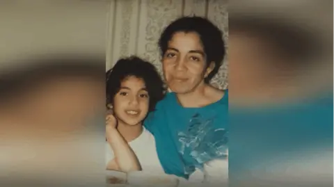 Nora Fakim/BBC A young girl sat next to a woman wearing a blue top. 