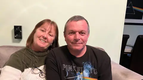 The image shows a middle-aged man and woman on a sofa. The woman is on the left and wearing an olive green and white jumper. She has spectacles on a chain around her neck. The man is on the right and wearing a Pink Floyd t-shirt
