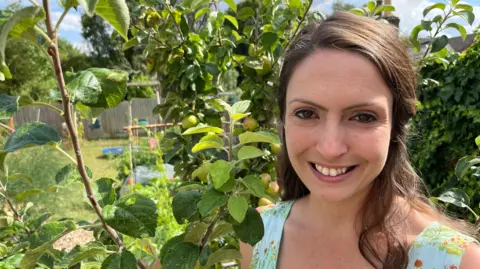 Alana Wilson is smiling at the camera. She is standing in a garden in front of an apple tree and children's toys can be seen in the background. She has long, dark hair and is wearing a flowery light blue top