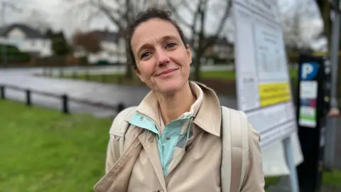 A woman with short dark hair and wearing a beige coat, looing straight at the camera. There's a grass verge, a road and some trees behind her.