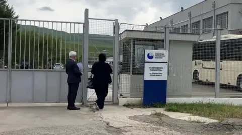 Rayhan Demytrie/BBC Two people, Malkhaz Salakaia and a female translator, can be seen entering a prison through a wire gate. Mr Salakaia is holding sheets of paper and wearing a suit.