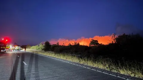 New Forest Police Fire in New Forest with orange glow seen to the right of a road and many emergency services in the distance.