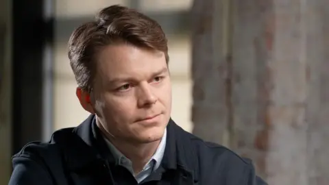 Palantir UK boss Louis Mosley speaking to the BBC. He is wearing a dark jacket with a button up jumper and is sitting in front of a grey wall. 