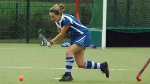 Clwb Hoci Rhuthun Hockey player Claerwen Render about to strike an orange ball with her hockey stick on an all weather pitch. She is wearing a blue hockey kit with blue and white striped socks.