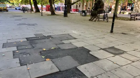 Carmelo Garcia A close-up of the 'tarmac cowpat' on Cheltenham's Promenade. A large patch of asphalt sticks out, surrounded by stone paving slabs. Shoppers can be seen walking in the distance. There are trees lining the Promenade, as well as benches and statues.