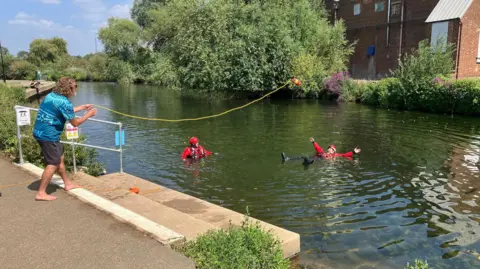 BBC/Sam Read A man is throwing a floatation ring toward two people dressed in red floating in a river