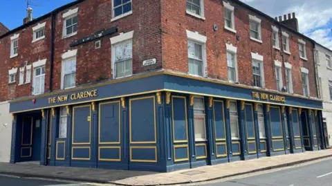 The New Clarence Building made of brick with a blue fascia. There are 22 windows on the first and second floors and four windows and two doors on the ground floor.