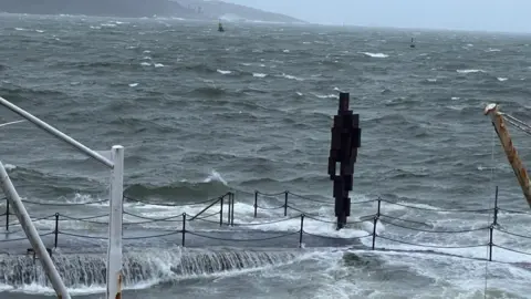 A wave crashes into a wall next to an Antony Gormley statue in Plymouth