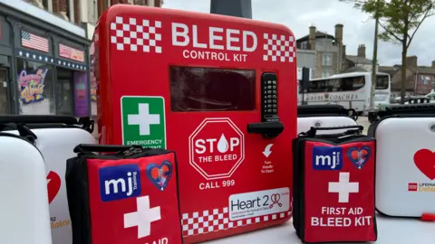 Richard Daniel/BBC A table loaded with the life-saving equipment, including white boxes containing defibrillators, red bleed kits and a large box protected by a pin pad that houses a bigger bleed kit.