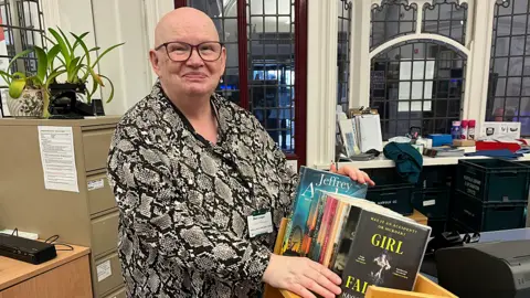 Librarian Mary Grogan stands by a shelf of colourful books waiting to be put back on the shelf. Mary is bald and wears brown glasses. She has a snake skin patterned shirt and wears her ID in a lanyard around her neck. 
