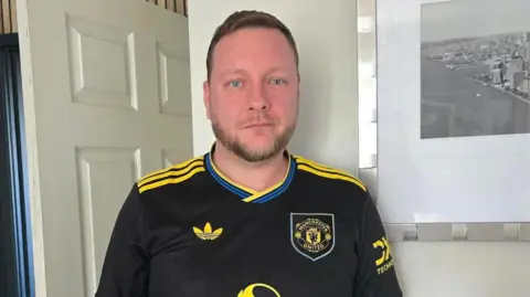 Hayley Coyle/BBC A man with short brown hair and beard wearing a black and yellow football shirt. He is standing in a cream-coloured living room