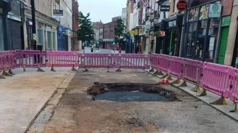 High Street in Bedford closed to traffic, with pink safety barriers surrounding a damaged section of road. Shopfronts line both sides of the street in the town centre.