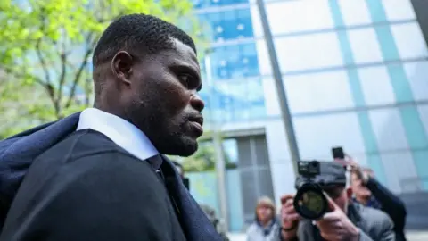 A black man with short hair in a black top with white collar protruding, is surrounded by photographers. The picture is taken side on and the man has a serious expression. 