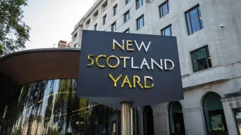 Sign for New Scotland Yard in front of a modern building with large windows and architectural details, under a clear sky.