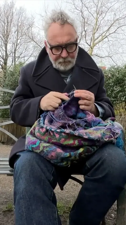 Elderly man with white hair and beard knitting on a bench outdoors