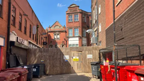 A temporary wooden fence with a danger sign on it sits across a courtyard, with a building behind missing its windows. There are waste bins in the foreground