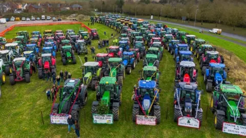 Devizes Young Farmers Around 150 tractors, decorated in tinsel and lined up in half a dozen neat rows on a field next to a road, ready to parade