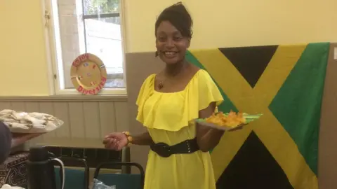 Cad Taylor/BBC A woman stands in a yellow dress in front of a Jamaican flag holding out a plate of food