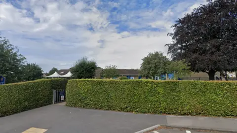 The exterior of a school. There are two large green hedges which are neatly trimmed and a small gate. There is a single-storey brick building behind the hedge. 