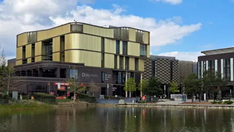BBC A large gold and black building that is several stories high and has large windows. There is a large pond not far from the building.