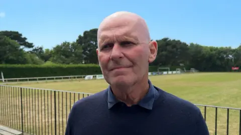A white man wearing a dark sweater and bald head. He is stood beside a fenced grass football field. In the distance you can see trees, a goalpost, and a clear blue sky.